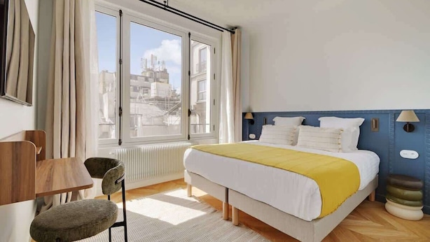 Une chambre d’hôtel équipée d’un lit, d’un bureau, d’une chaise et offrant une vue sur la ville grâce à de grandes fenêtres.