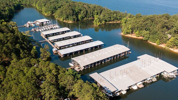 An aerial view of a marina with multiple docked boats and covered boat storage areas surrounded by dense forest.