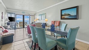 A spacious living area with a glass dining table, teal chairs, and a ceiling fan.