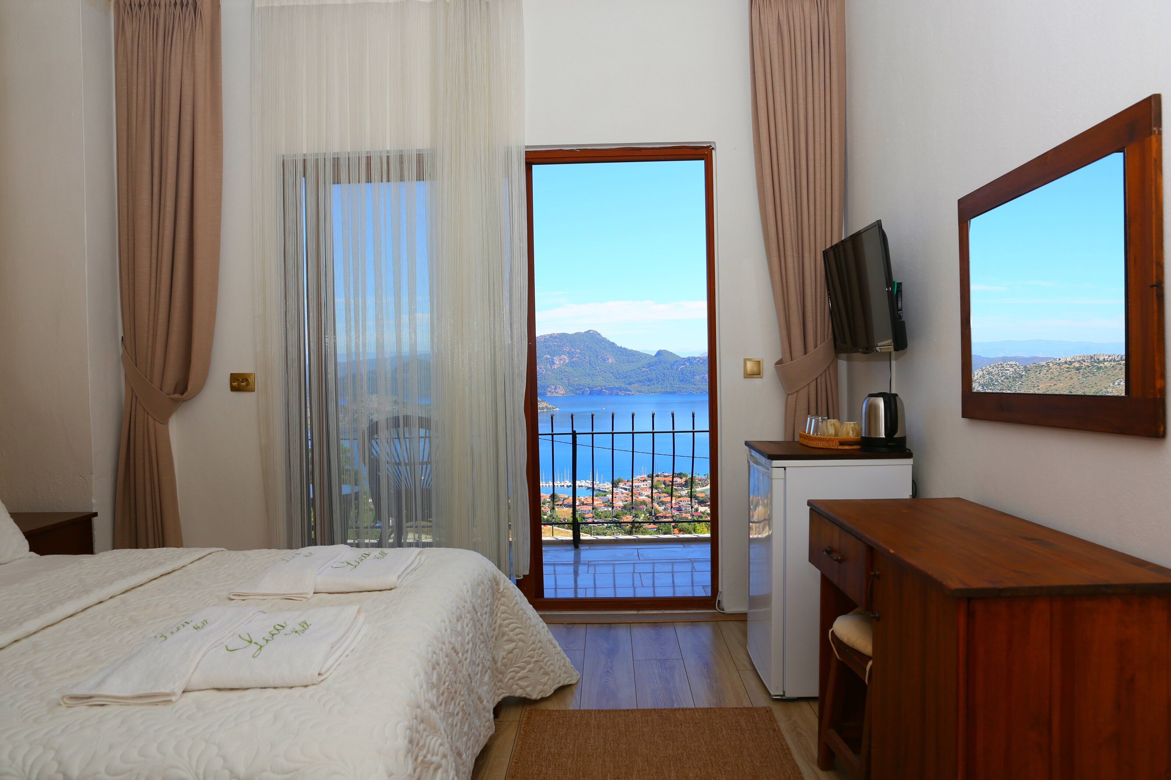 Habitación de hotel con cama, escritorio, televisión y balcón con vistas al mar y a las montañas.