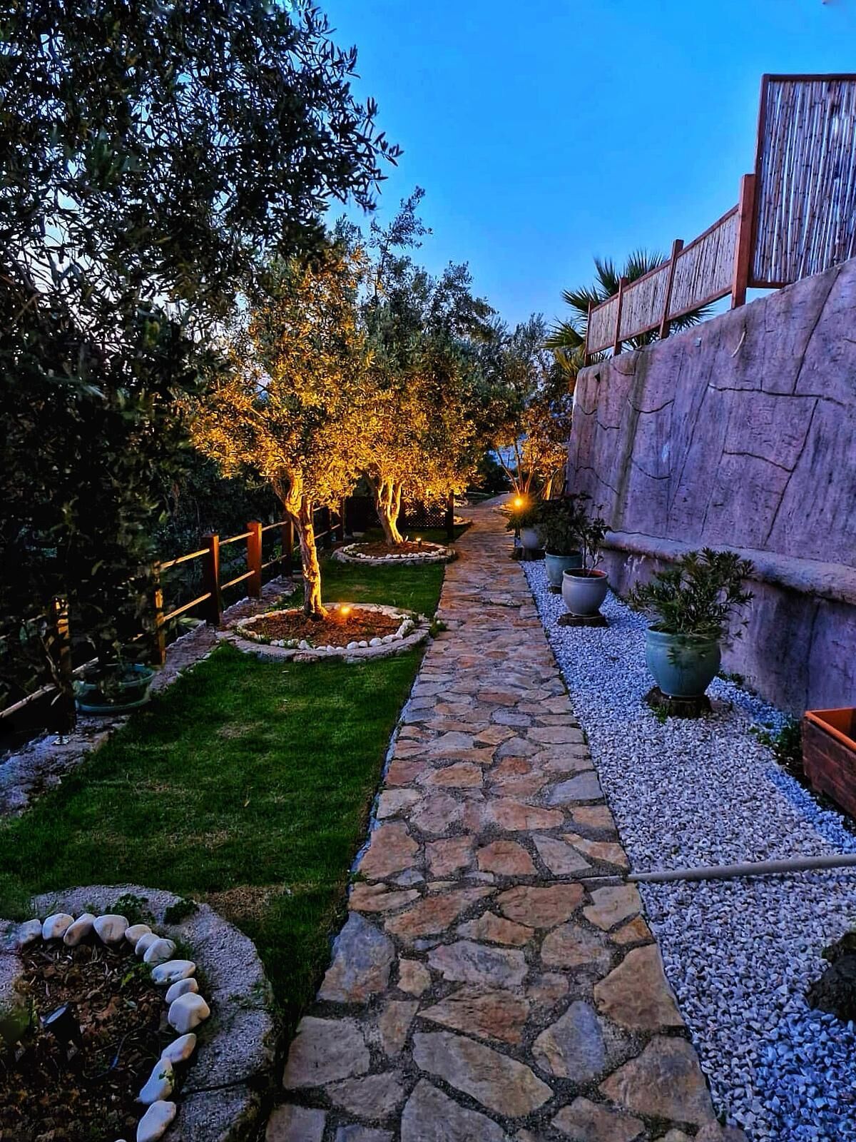 A stone pathway leading through a garden with trees and a rock wall on one side.