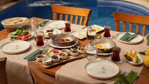 A dining table set for a meal with various dishes and drinks, surrounded by chairs and an umbrella, with a pool in the background. - Loca Hill (Marmaris)