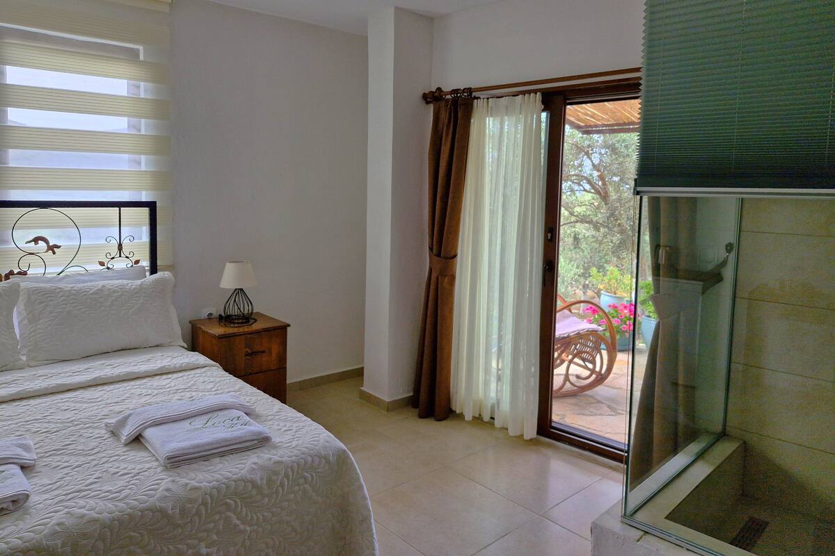 A hotel room with a bed, a nightstand, a lamp, a window with blinds, and a sliding glass door leading to an outdoor area with a chair and potted plants.