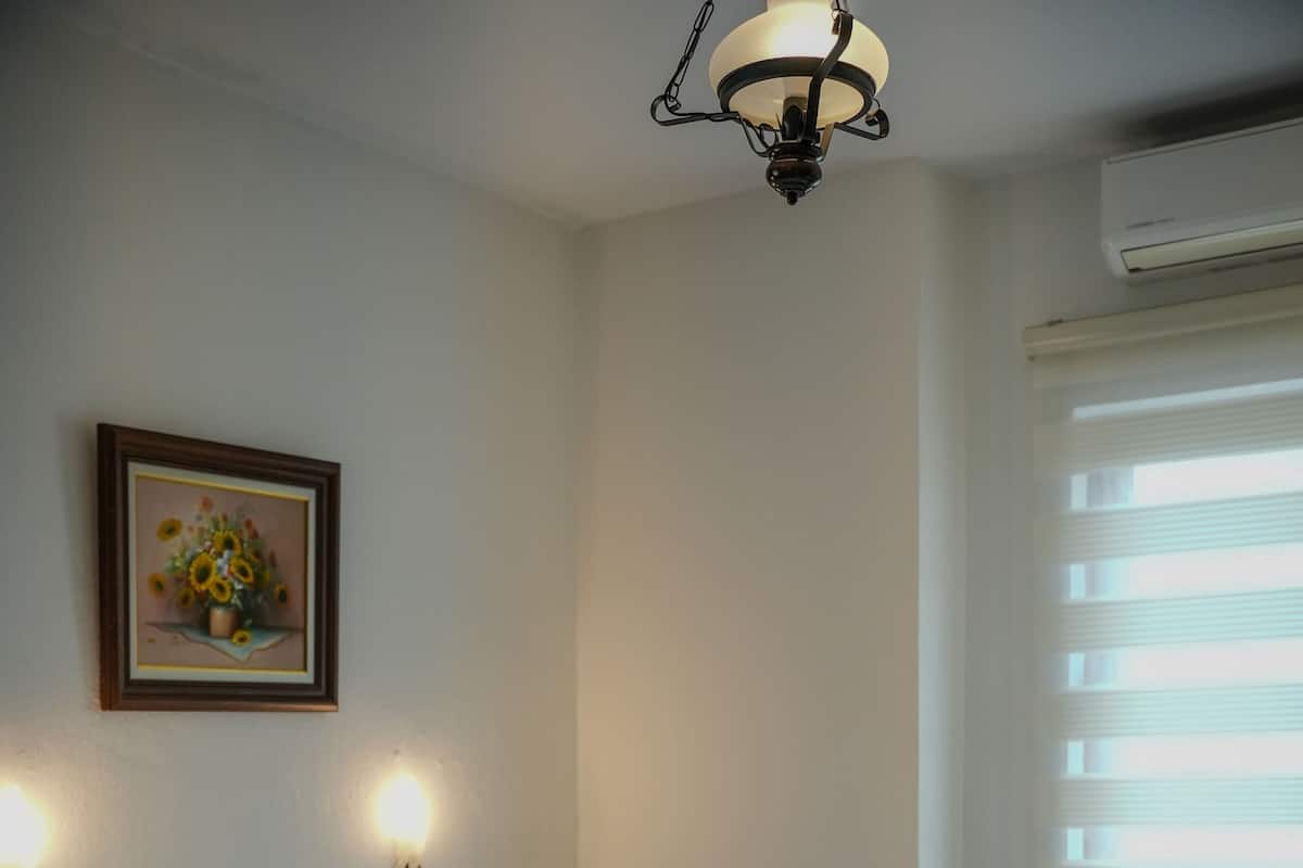 A bedroom with a wooden bed, white bedding, two wall-mounted lights, a painting, and a window with blinds.