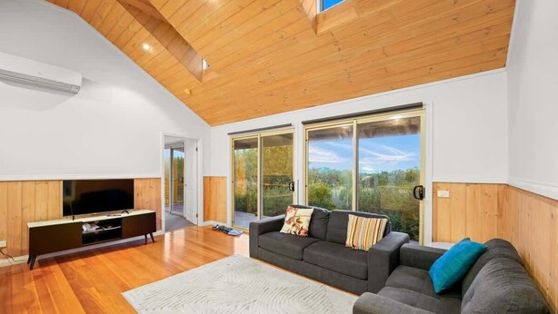 A modern living room with a wooden ceiling, a flat-screen TV, a gray sofa, and a large window offering a view of the outdoors.