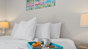 Una cama bien hecha con ropa de cama blanca, una bandeja azul con pasteles y dos tazas blancas, y una pared con tres cuadros de flamencos.