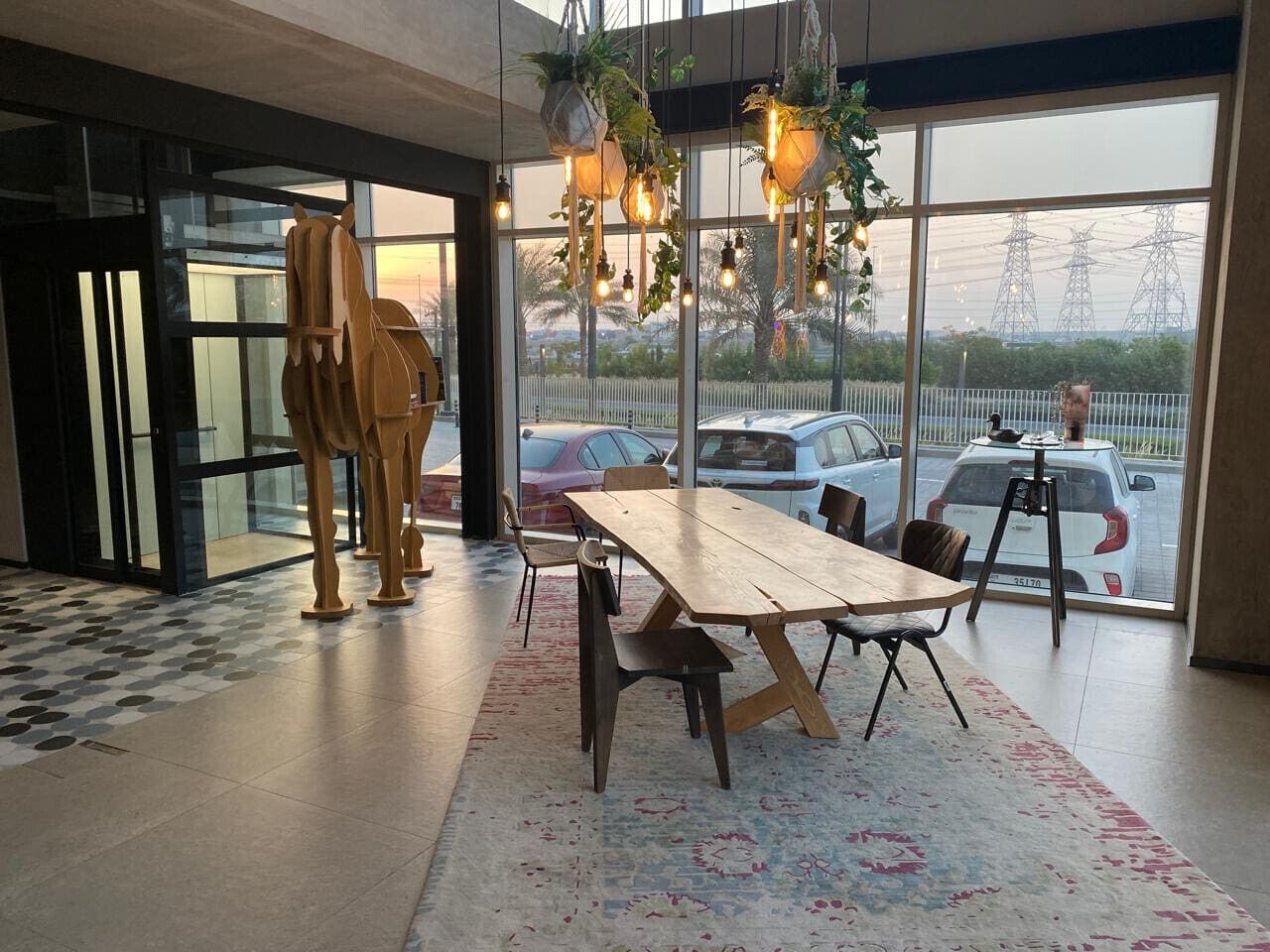 Um interior moderno com uma mesa e cadeiras de madeira, grandes janelas com vista para carros estacionados e linhas de energia, e vasos suspensos com luzes.