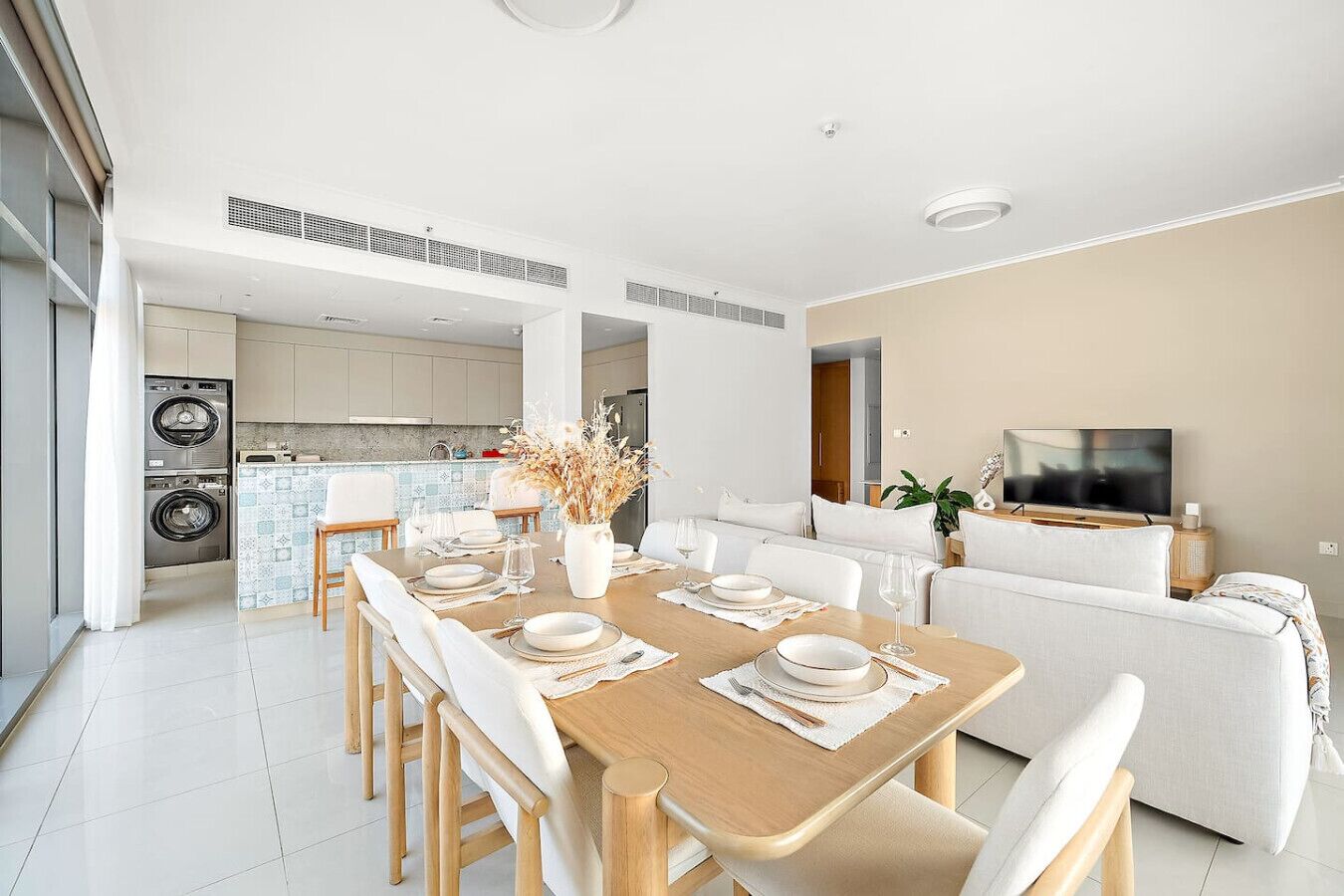 Uma cozinha moderna com eletrodomésticos embutidos, uma sala de jantar com mesa de madeira posta para quatro pessoas e uma sala de estar com sofá e televisão.