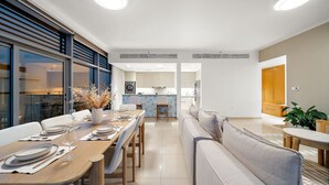 A modern dining area with a long table set for four, a sofa, and a kitchen in the background.