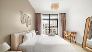 A modern hotel room with a large bed, a desk, a chair, a mirror, and a view of the cityscape through the window.