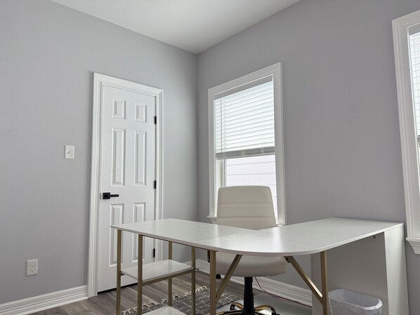 A room with a white desk, a chair, a window with blinds, a door, and a small rug.