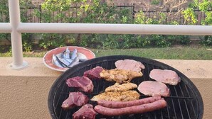 En grill med olika sorters kött och fisk står på en balkong med utsikt över en park och träd.