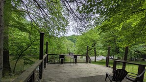 A wooden deck with chairs and a table overlooking a river, surrounded by dense green trees.