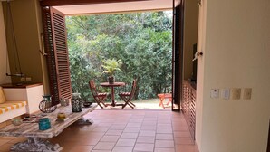 Una stanza con pavimento piastrellato, una panca di legno, un tavolino con sedie e vista su un giardino rigoglioso attraverso una porta aperta.