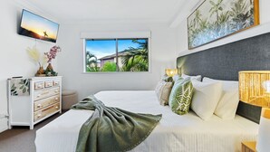 A bedroom with a bed, a TV, a window with a view of palm trees, a dresser, and a painting on the wall. - Family Friendly 2BR Apartment with Resort Pool (Westcourt)