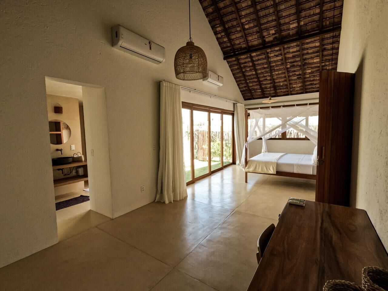 Une pièce spacieuse avec une table en bois, un luminaire suspendu, un lit à baldaquin et une vue sur l’extérieur grâce à de grandes fenêtres.