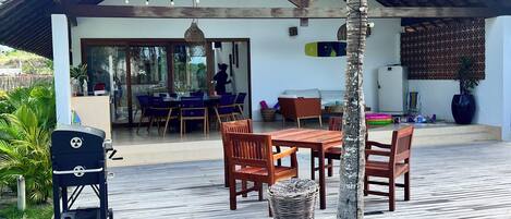A covered outdoor patio with wooden furniture, a grill, and a palm tree.