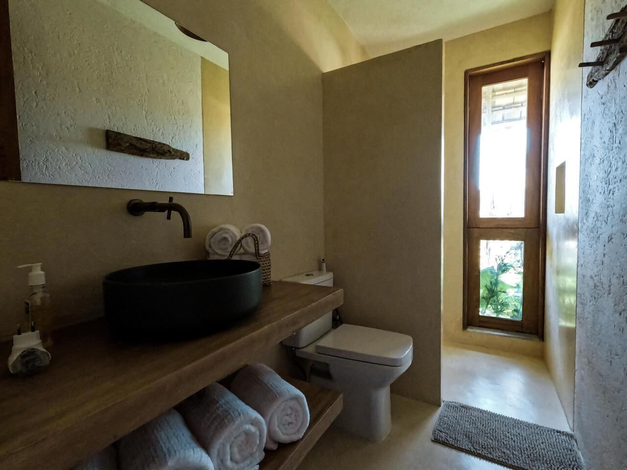 A bathroom with a wooden vanity, black sink, white toilet, and a window with a view of greenery.
