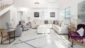 A spacious living room with a large sectional sofa, a round coffee table, and a purple armchair. There is a chandelier hanging from the ceiling and artwork on the walls. - 5 Bedroom Home With Firepit (San Diego)