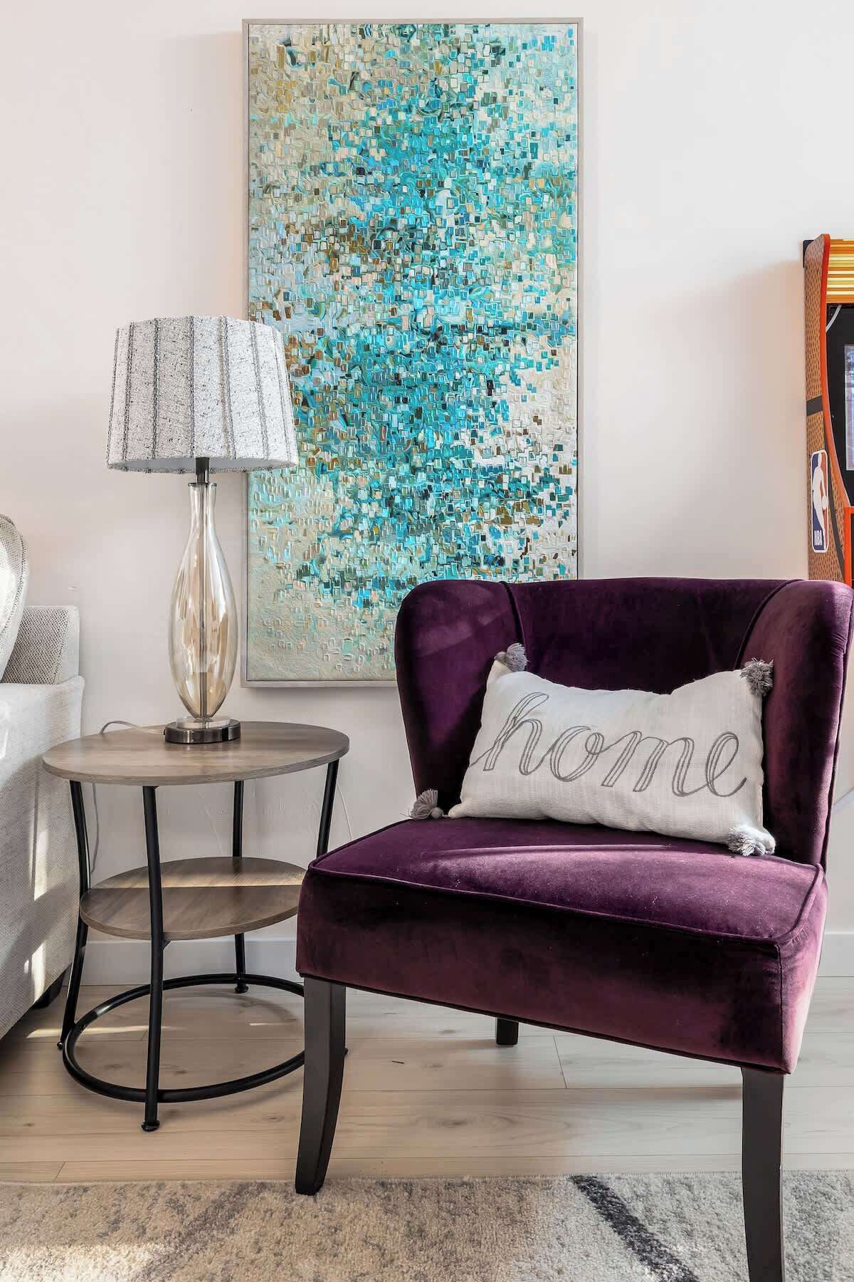 A living room with a purple velvet armchair, a round side table with a lamp, and a large abstract painting on the wall.