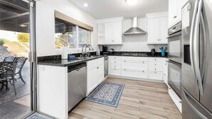A modern kitchen with white cabinets, stainless steel appliances, and a large window overlooking a patio. - 5 Bedroom Home With Firepit (San Diego)
