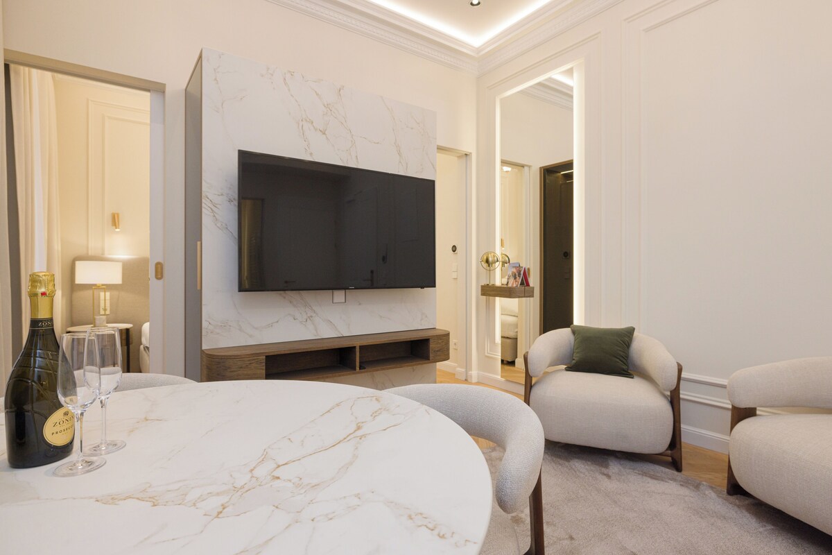 A modern living room with a marble fireplace, a flat-screen TV mounted above it, and a round dining table with wine glasses.