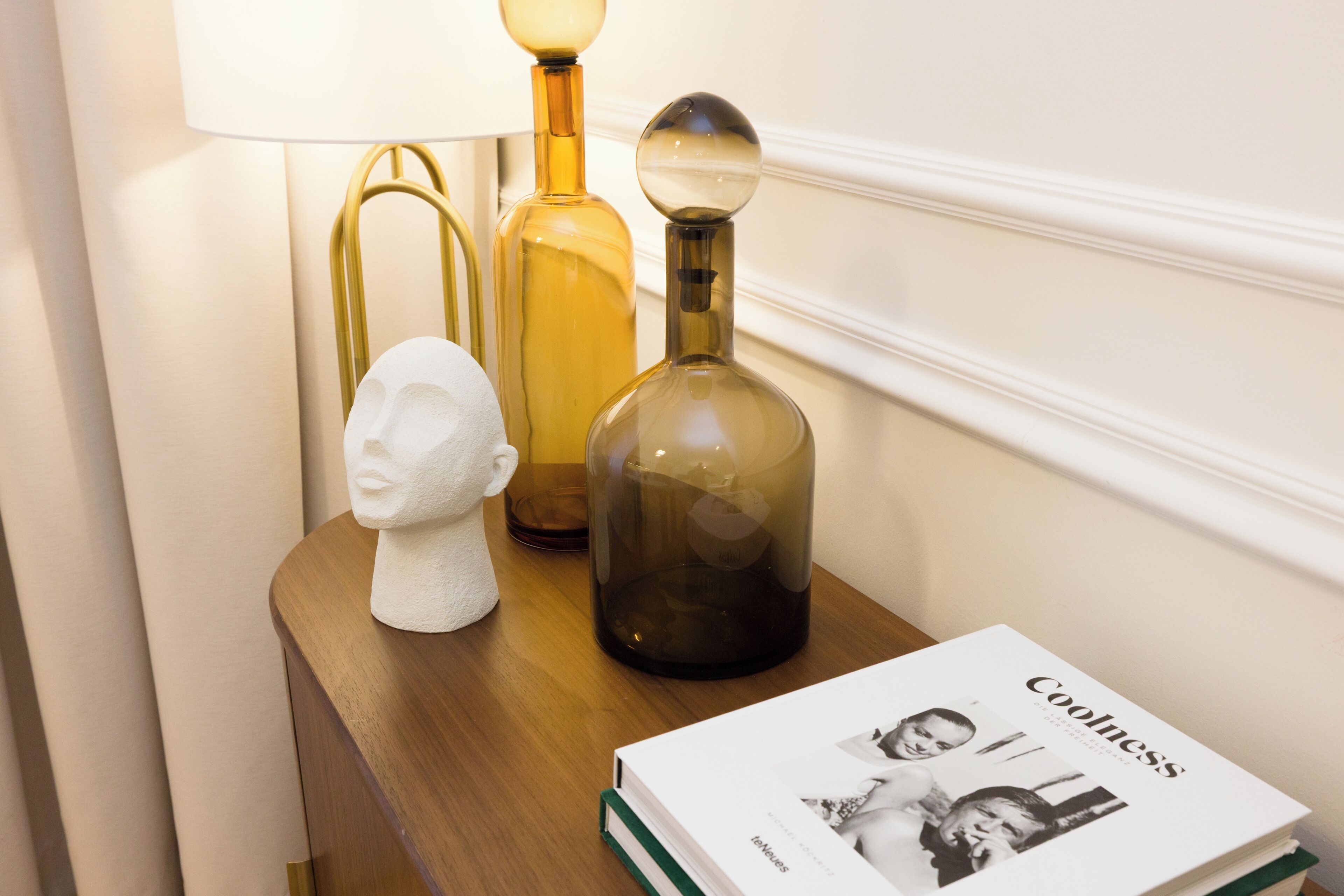 A wooden side table with a white bust, a lamp, and a book titled 'Coolness' on a wall with a decorative molding.