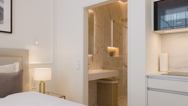 A hotel room with a bed, bedside lamp, a bathroom with a marble wall, a sink, and a television mounted above the sink. - RAFAEL KAISER - Signature Apartments  (Vienna)