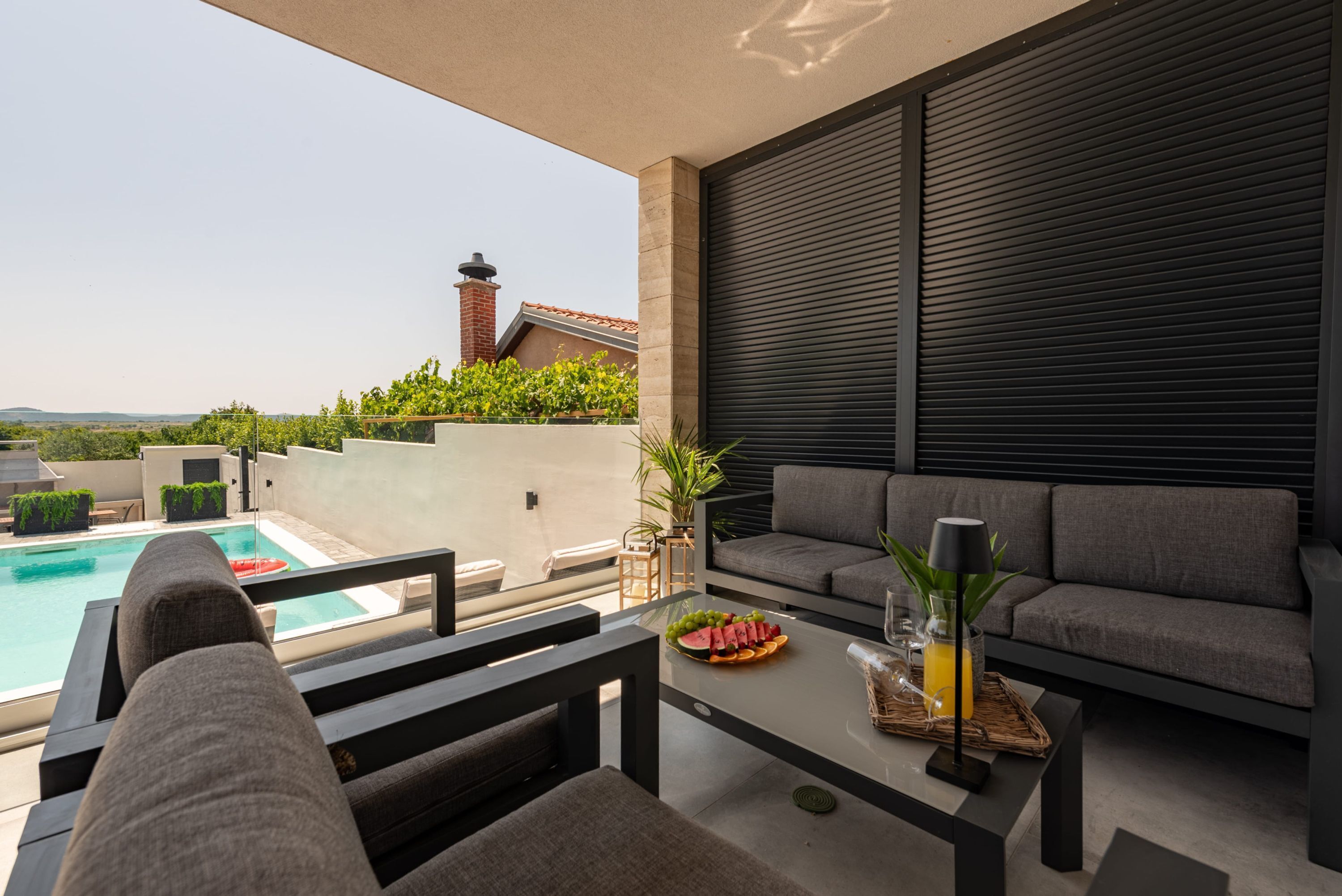 A modern outdoor seating area with a pool, a dining table with fruit and drinks, and a view of a house with a chimney.
