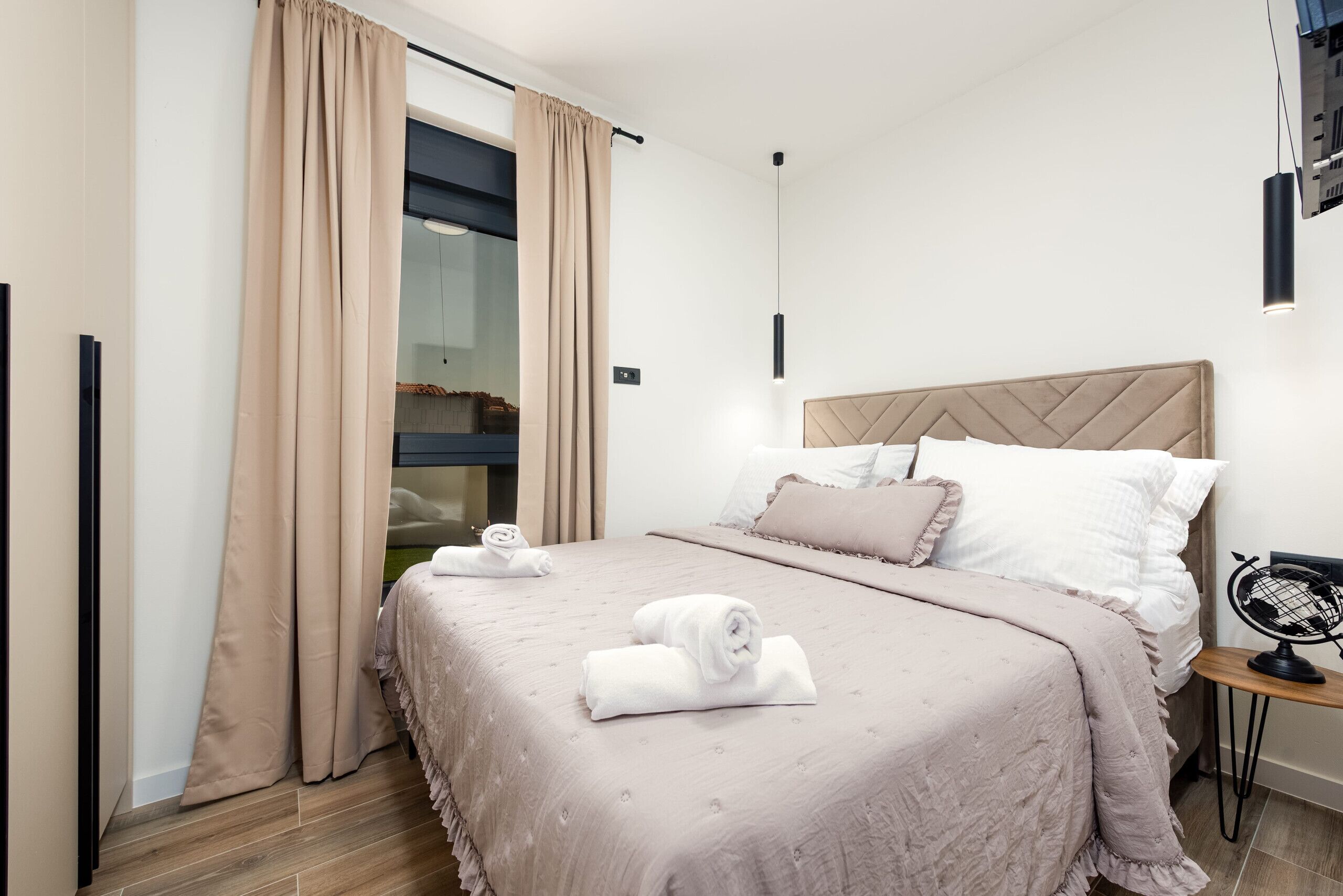 Une chambre moderne avec un grand lit, des rideaux beiges, une petite table sur laquelle est posé un globe terrestre et un luminaire suspendu.
