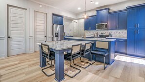 A modern kitchen with blue cabinets, a stainless steel refrigerator, a microwave, and a dining area with a glass-top table and chairs.