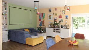 A living room with a sofa, ottoman, dining table with fruit, and a kitchen area with cabinets and a window.