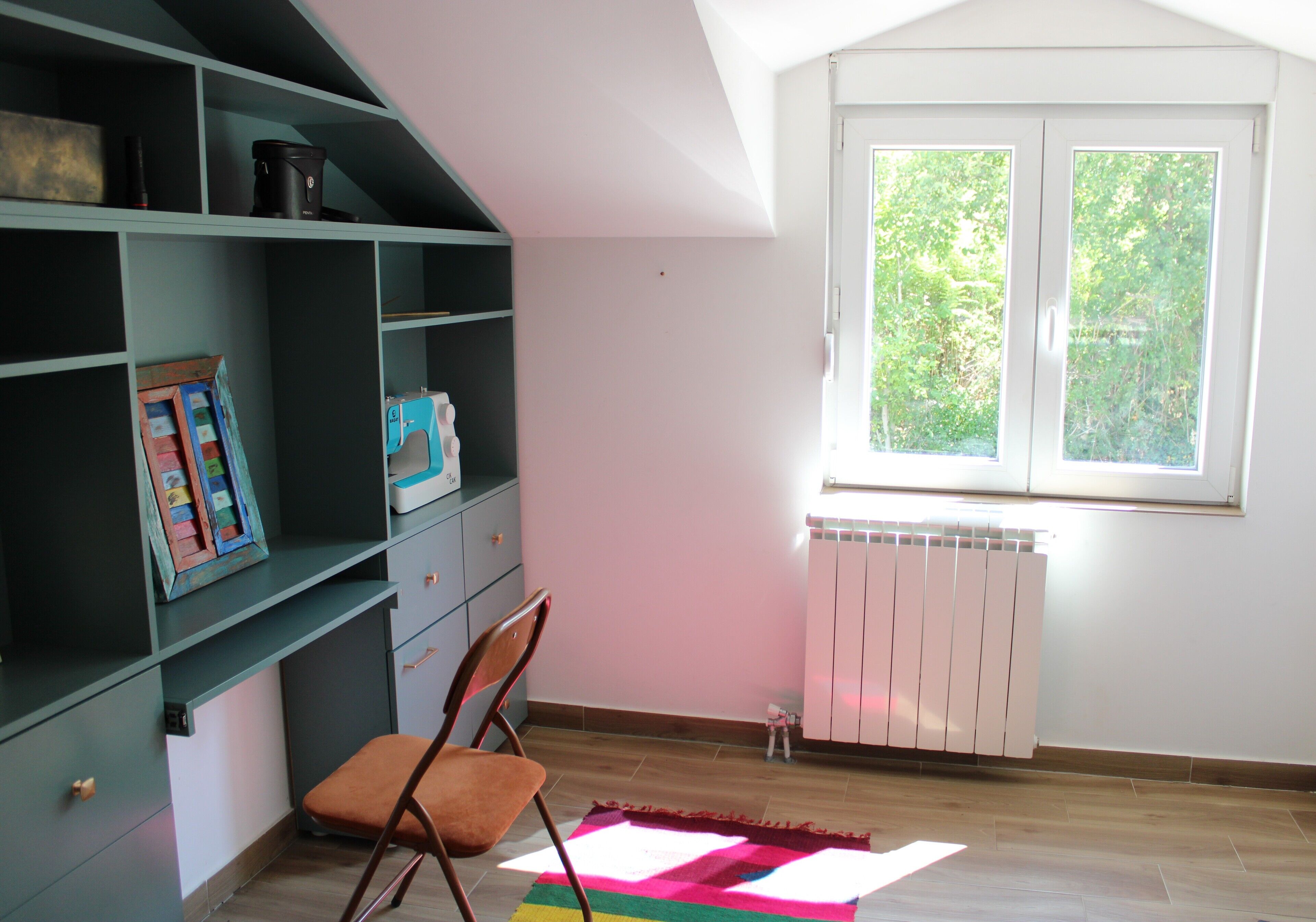 A room with a sloped ceiling, a window with a view of greenery, a radiator, and a chair with a colorful rug.