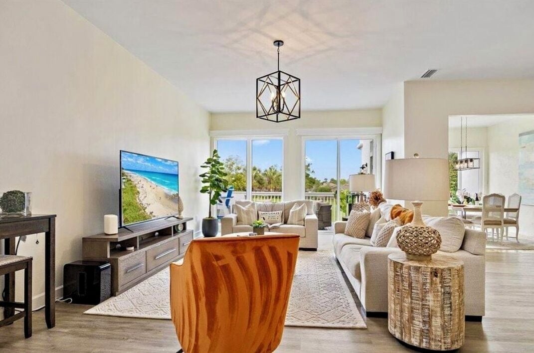 A modern living room with a large flat-screen TV, a wooden accent chair, a round wooden side table, and a beige sofa with multiple cushions.
