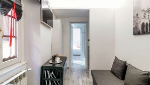 Un couloir étroit avec un radiateur, un meuble noir sur lequel se trouve un plateau rempli d’objets, un canapé gris et un tableau encadré accroché au mur.