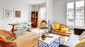 A bright living room with a wooden floor, a sofa with yellow cushions, a dining table with chairs, and a large window with a view of buildings.
