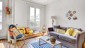 A cozy living room with a blue-striped rug, a grey sofa with yellow pillows, a white armchair with colorful cushions, and a hanging plant.