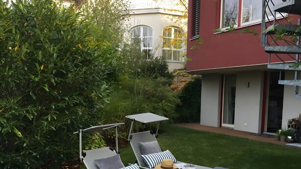 A garden with two lounge chairs and a small table, surrounded by greenery and a building with large windows. - Room 'Hortensie' with Private Terrace, Private Garden and Wi-Fi (Weil der Stadt)