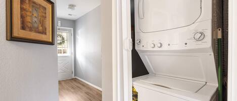 Una lavandería con lavadora y secadora blancas, un cuadro enmarcado en la pared y una puerta que da a un pasillo con piso de madera.
