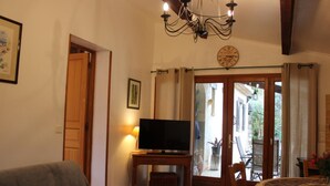 A cozy living room with a television on a wooden stand, a sofa, a dining table with chairs, and a view of the outside through a glass door.