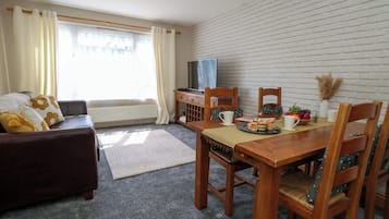 Un salón acogedor con un sofá marrón, una mesa de comedor de madera dispuesta con platos, tazas y un cuenco de fruta. Un televisor está sobre un mueble de madera, y se ve una pared de ladrillo blanco.