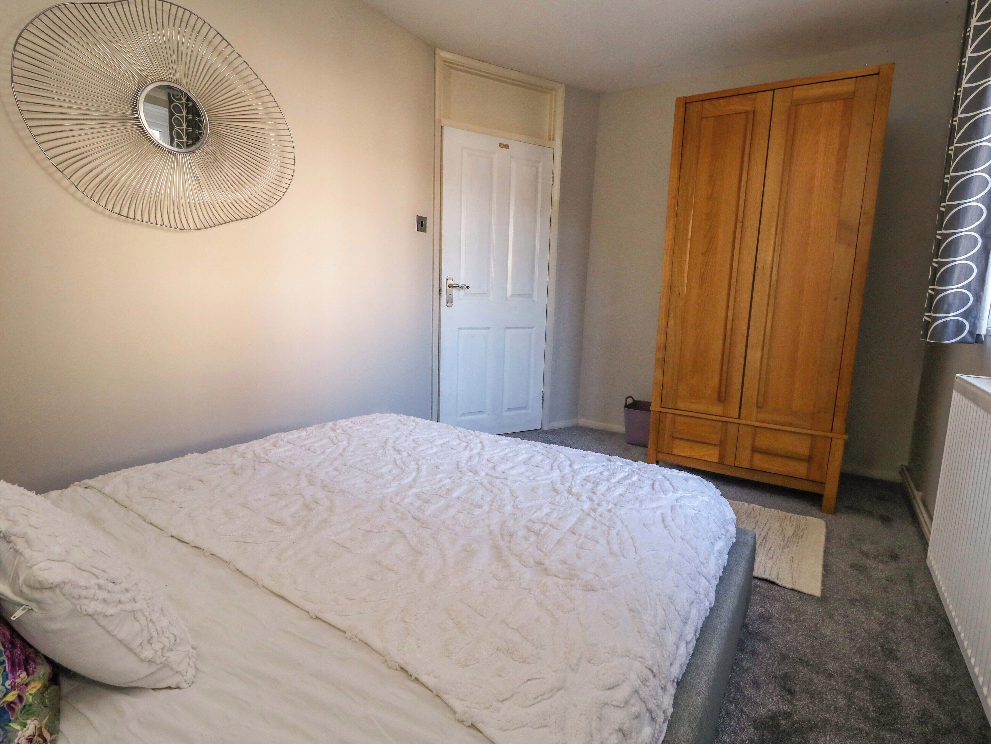 A bedroom with a large bed, a wooden wardrobe, a white door, and a wall-mounted mirror.