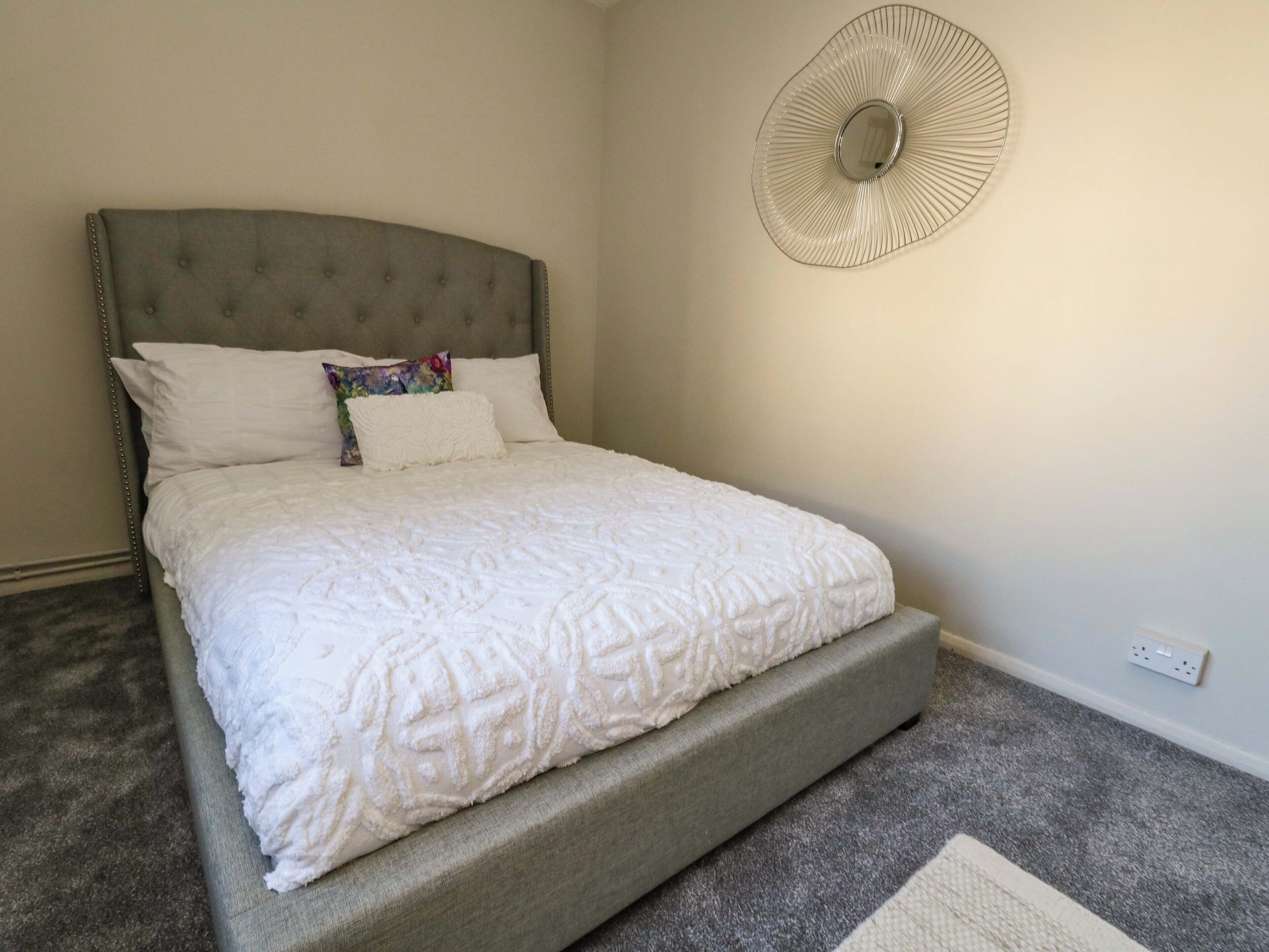 Cama bem arrumada com cabeceira estofada, um leque decorativo na parede e piso coberto por carpete.