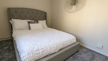 Cama bem arrumada com cabeceira estofada, um leque decorativo na parede e piso coberto por carpete.