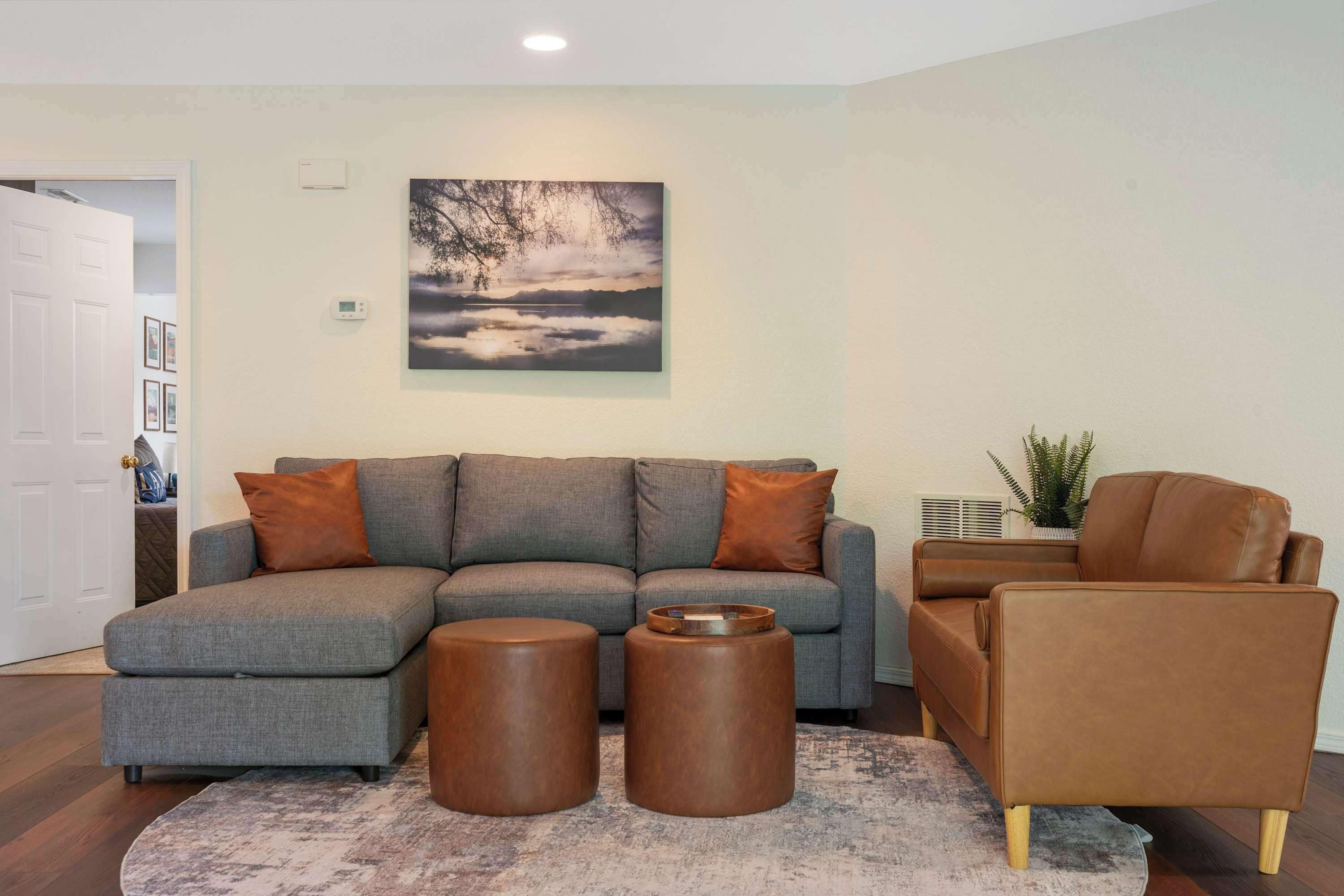 Una sala de estar con un sofá seccional gris, dos otomanas marrones, un sillón de cuero marrón y un cuadro de un árbol en la pared.