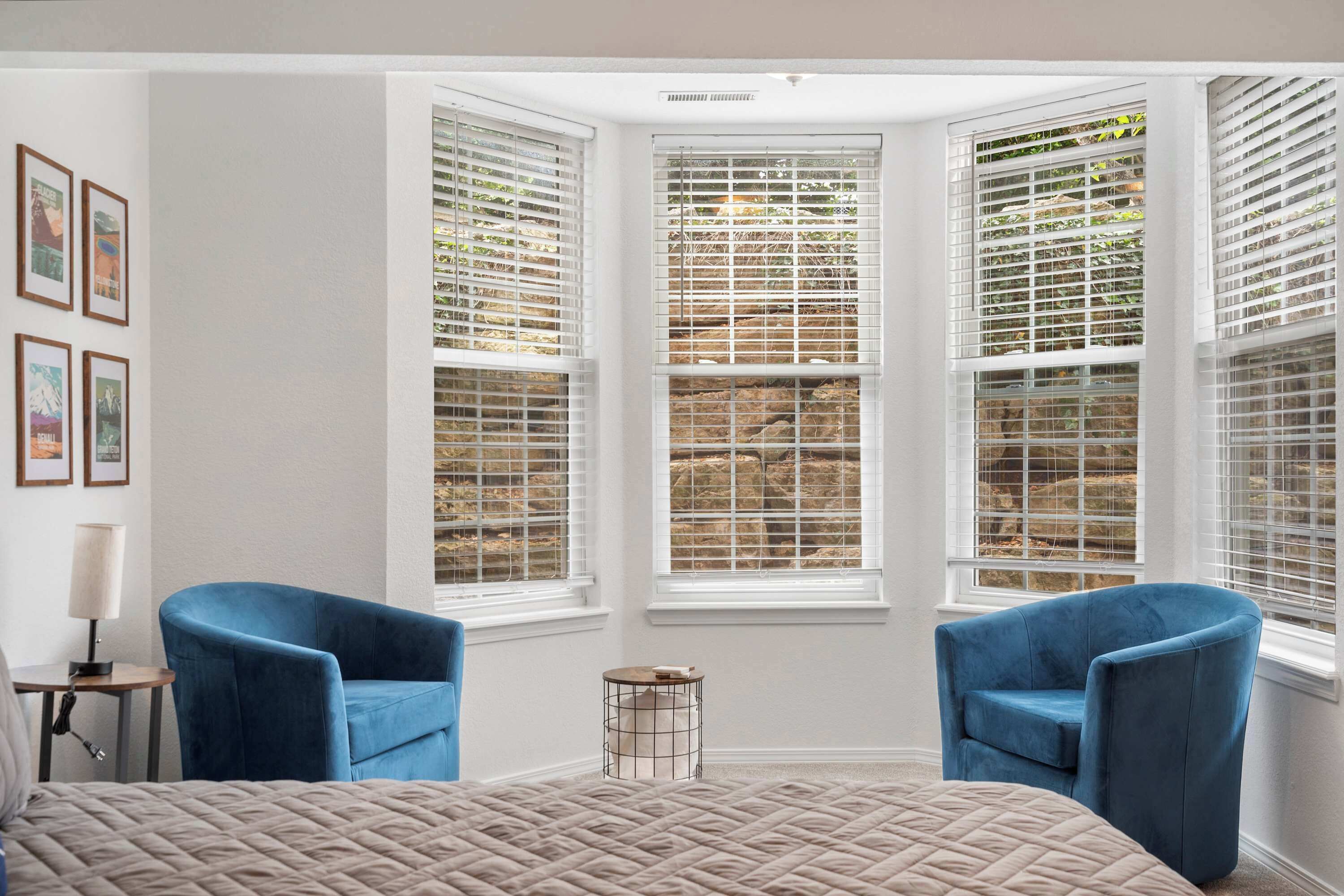 A room with a large bed, two blue chairs, a small round table, and a window with blinds.