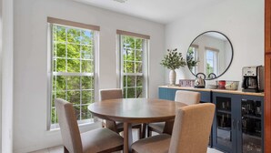 A dining area with a round table and four chairs, large windows with a view of trees, a kitchen area with cabinets and a coffee maker, and a round mirror on the wall. - Lake It Easy Condo for 7 (Kimberling City)