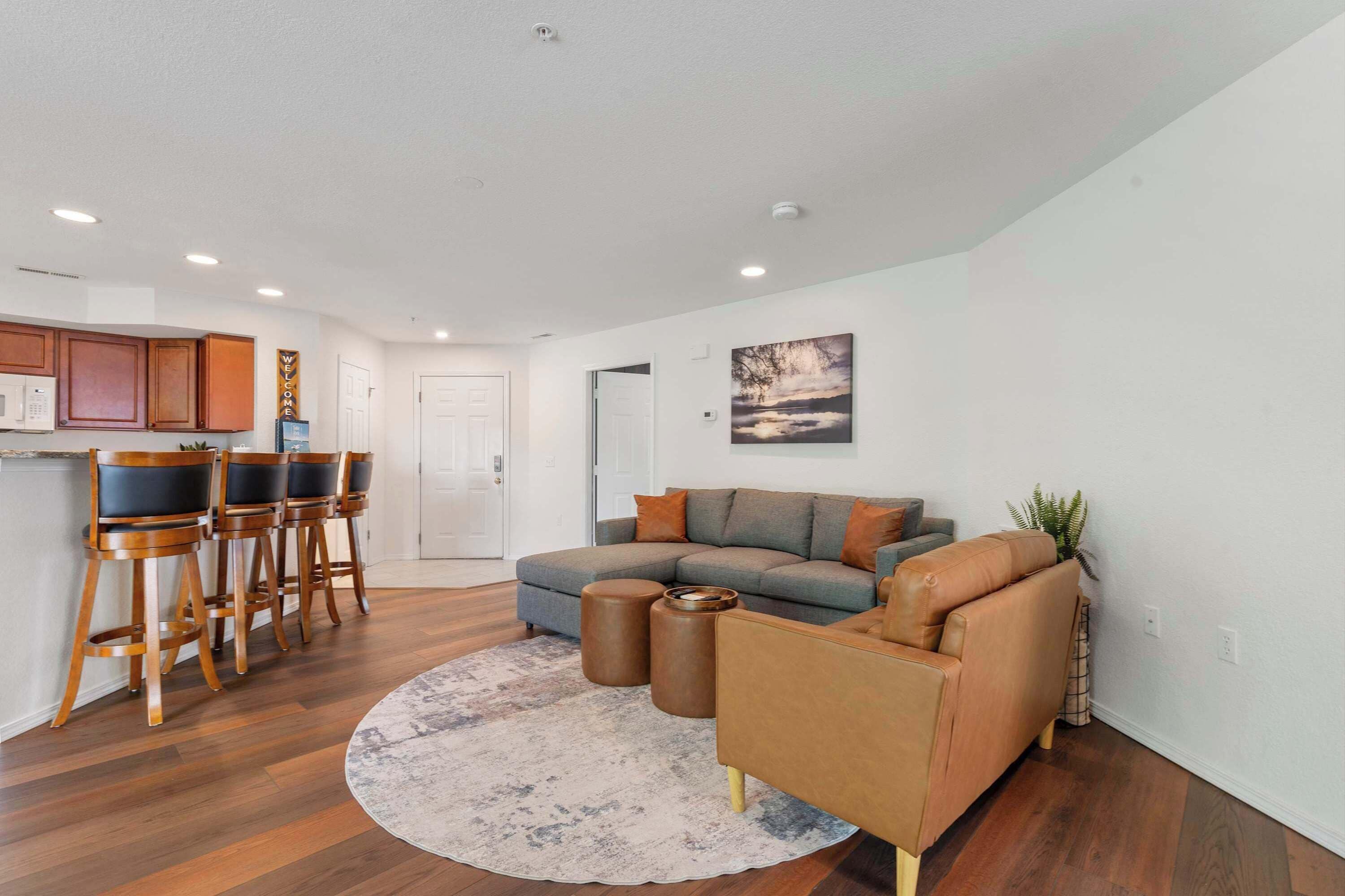 A modern living room with a grey sofa, brown leather armchair, and wooden bar stools.