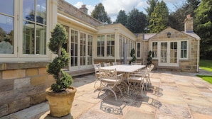 A patio with a table and chairs, a potted plant, and a stone building with large windows.