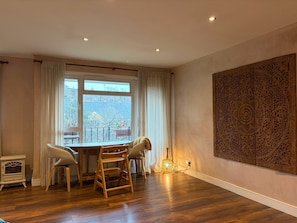 A room with a wooden floor, a dining table with chairs, a large window with curtains, a wall-mounted artwork, and a small table with a lamp.